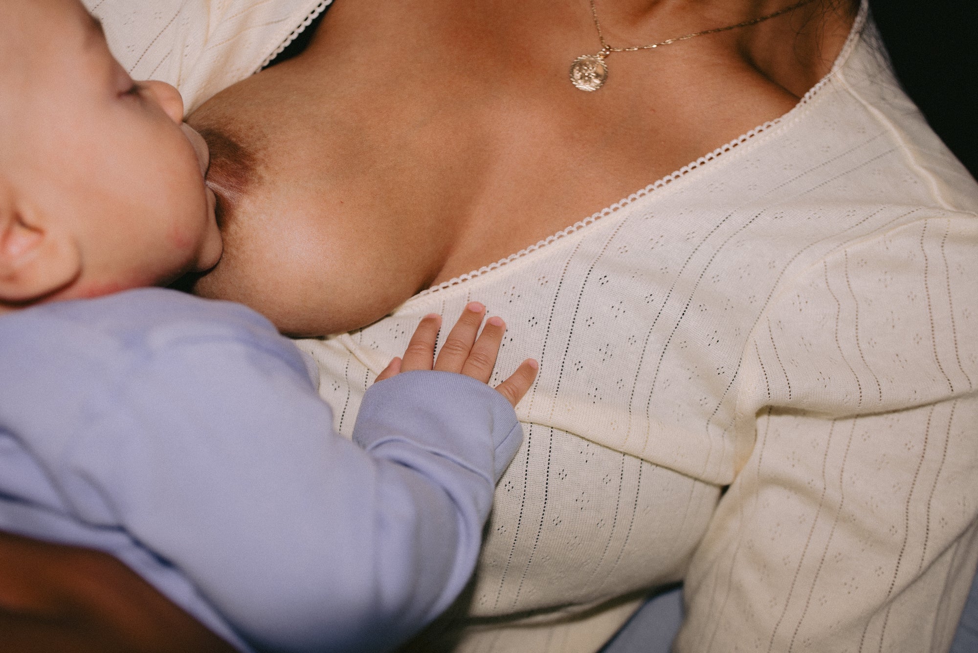 Close-up of a mother breastfeeding her baby boy in a blue onesie, with his hand resting gently on her chest and a gold necklace visible.