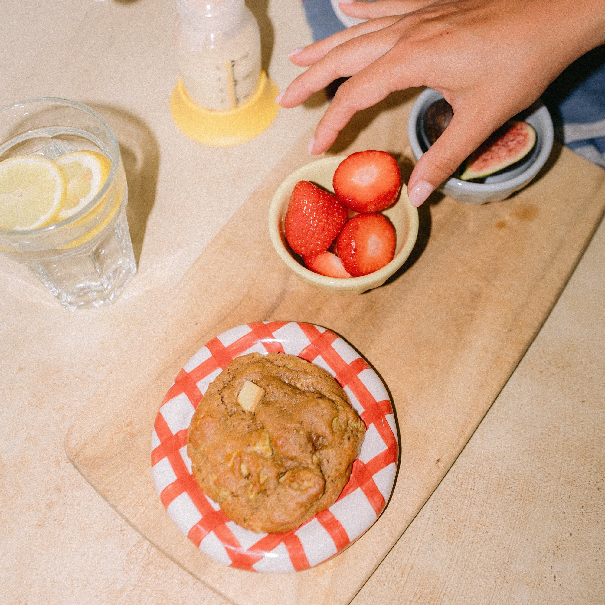 Chai spiced lactation cookie with pistachio and white chocolate, on a kitchen counter with lemon water, breast pump, and a mum reaching for a strawberry