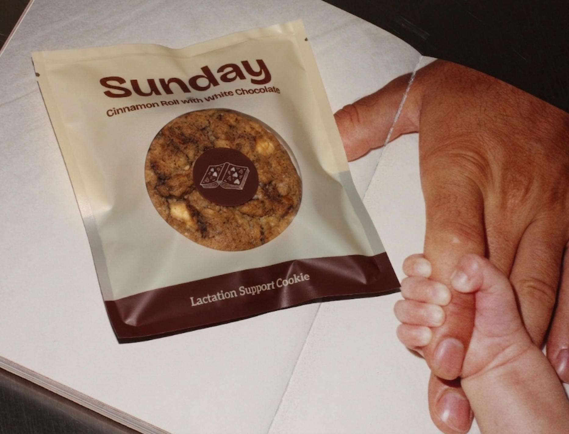A person holding a baby's hand next to a package of a Sunday Lactation Support Cookie. The package is cream-colored with a dark brown bottom banner.
