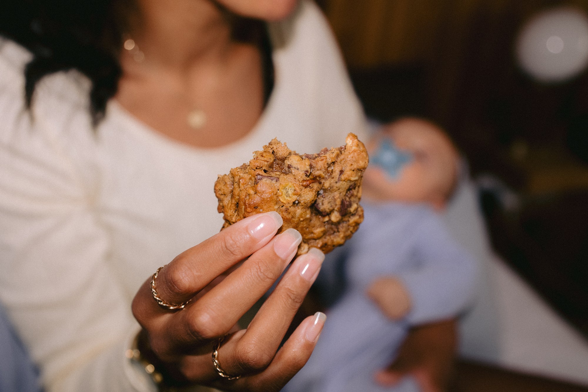 Cowgirl lactation cookie with pecan, coconut, cornflakes, and dark chocolate, held by a mum wearing gold rings while holding her baby in the other arm