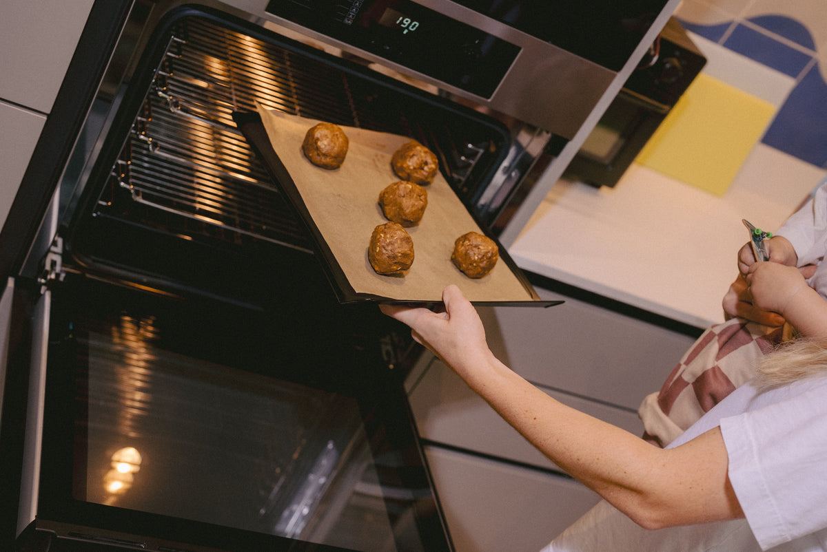 A mother holding her child in a white shirt places a baking tray with six lactation support oatmeal chocolate chip cookie dough balls into a black built-in oven inside a modern kitchen.
