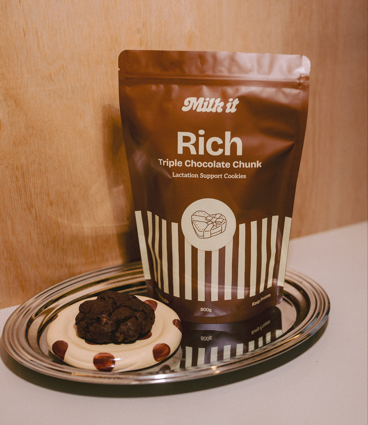 A stand-up pouch of Milk It Rich Triple Chocolate Chunk lactation support cookies displayed on a reflective silver tray next to a single triple chocolate cookie, set against a warm wood-paneled background.