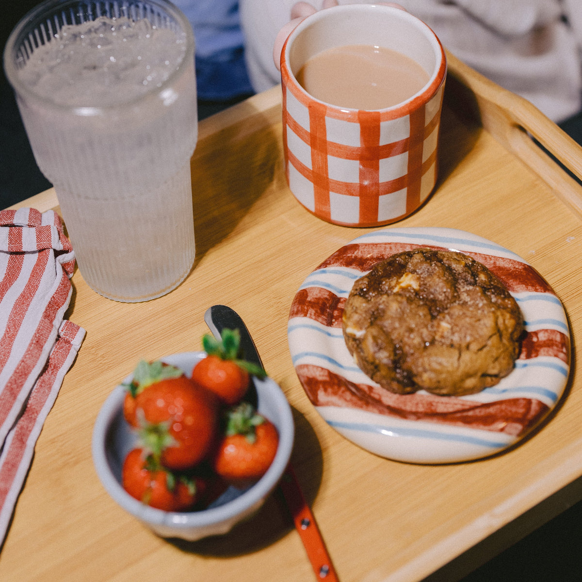 A cosy snack scene on a wooden tray with a striped mug filled with tea, a glass of iced water, strawberries in a small bowl, and a lactation support cookie on a striped plate. A person in a blue denim jacket and white sweater is seated in the background.