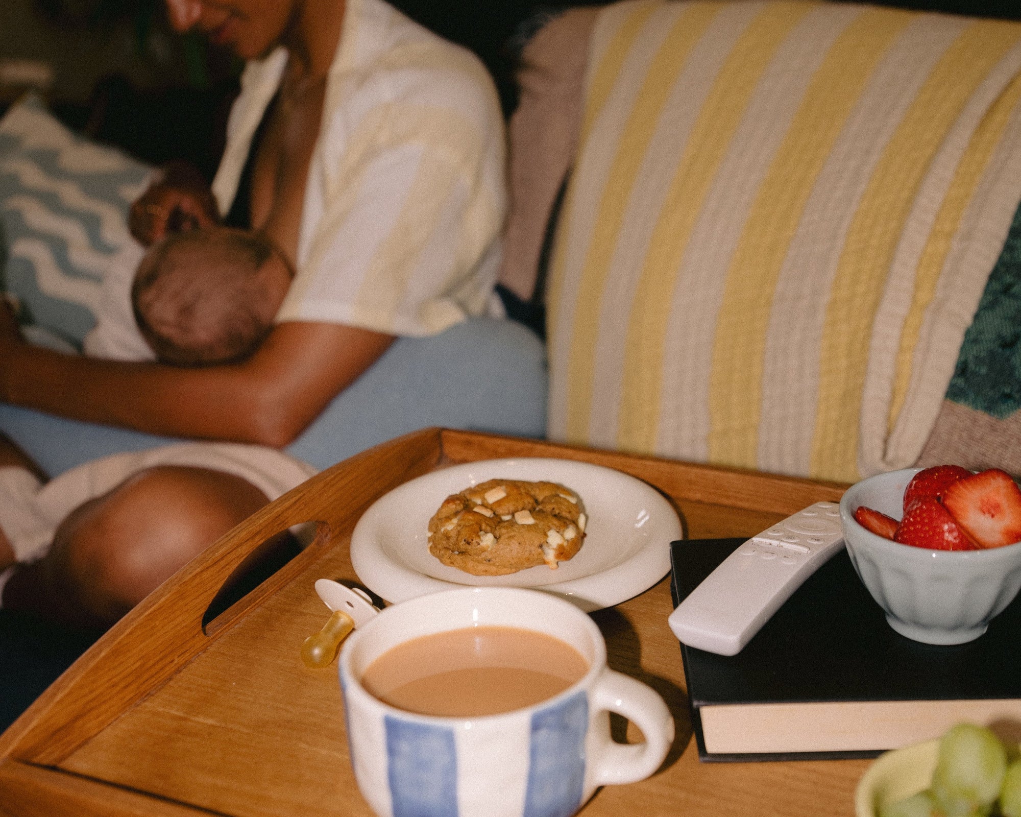 A comforting postpartum scene featuring a Milk it lactation cookie on a tray with a cup of tea, fresh strawberries, a remote, and a book — with a mum breastfeeding in the background. A quiet moment of nourishment and care.