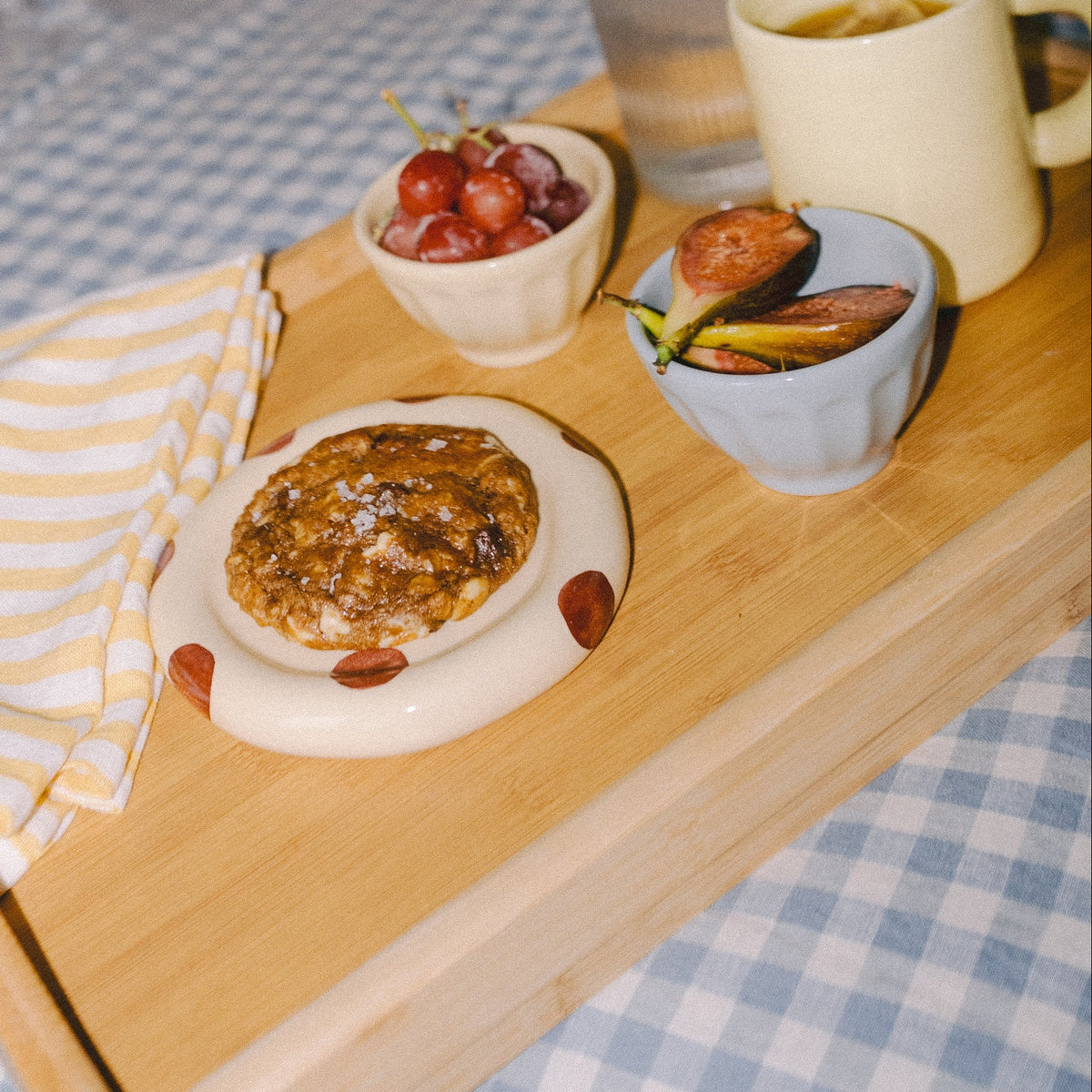 A wooden breakfast tray with lactation support cookies and a mug sits on a blue-and-white checkered bed in a bedroom, with a woman holding a baby in the background.