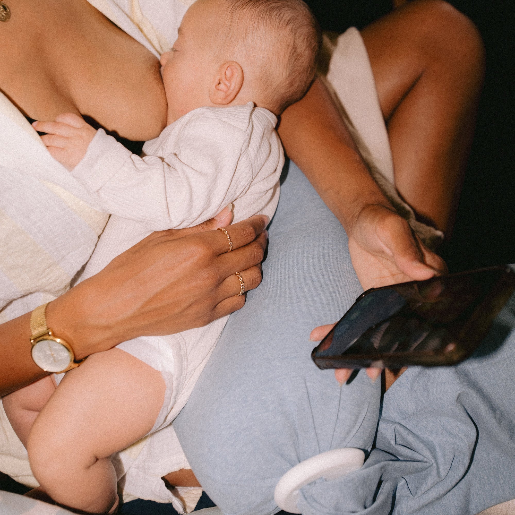 Breastfeeding mother holding baby while she breastfeeds using a breastfeeding pillow while scrolling on her phone