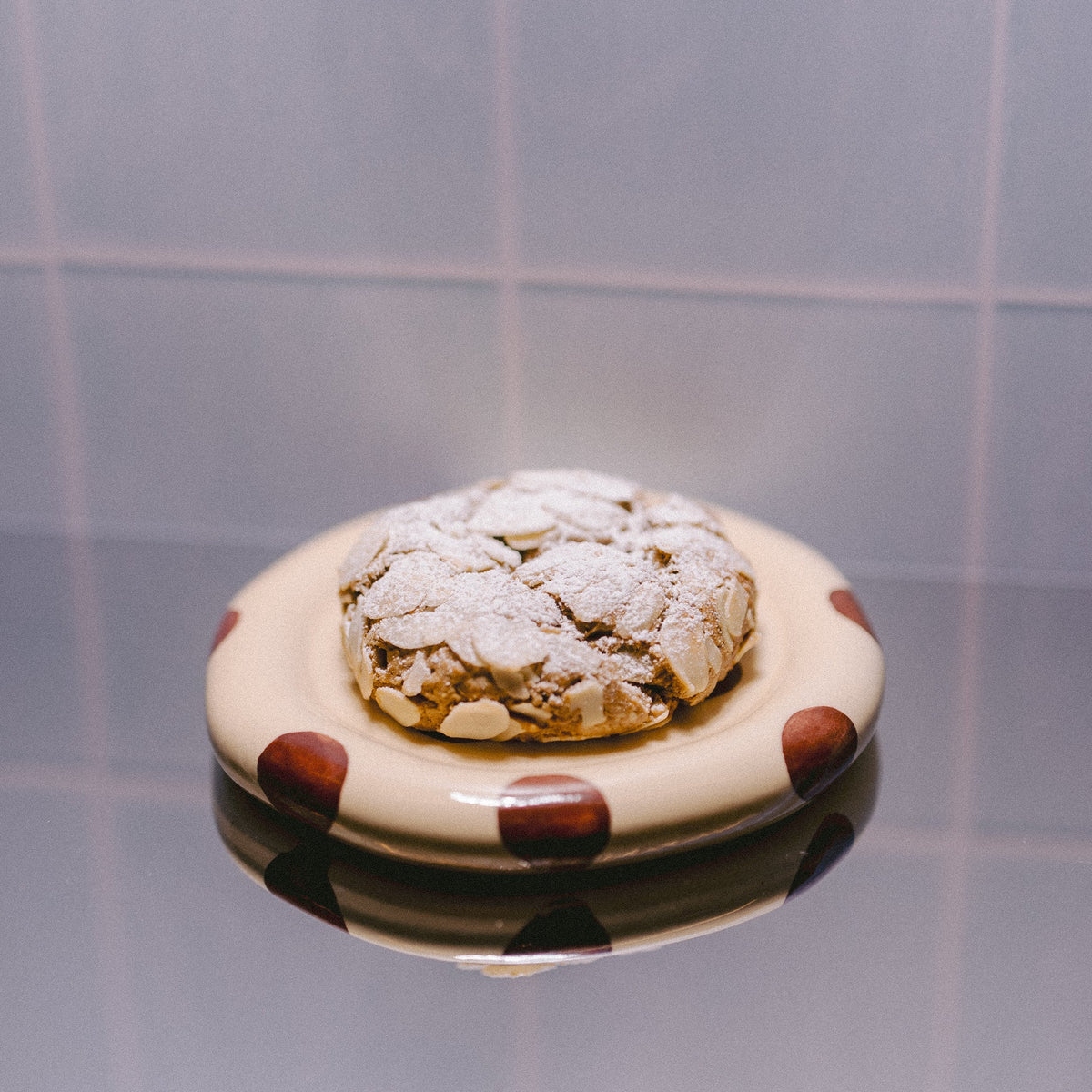 A small round plate with a toasted almond croissant lactation support cookie with powdered sugar on top of the cookie, set against a tiled background with a reflective surface below.