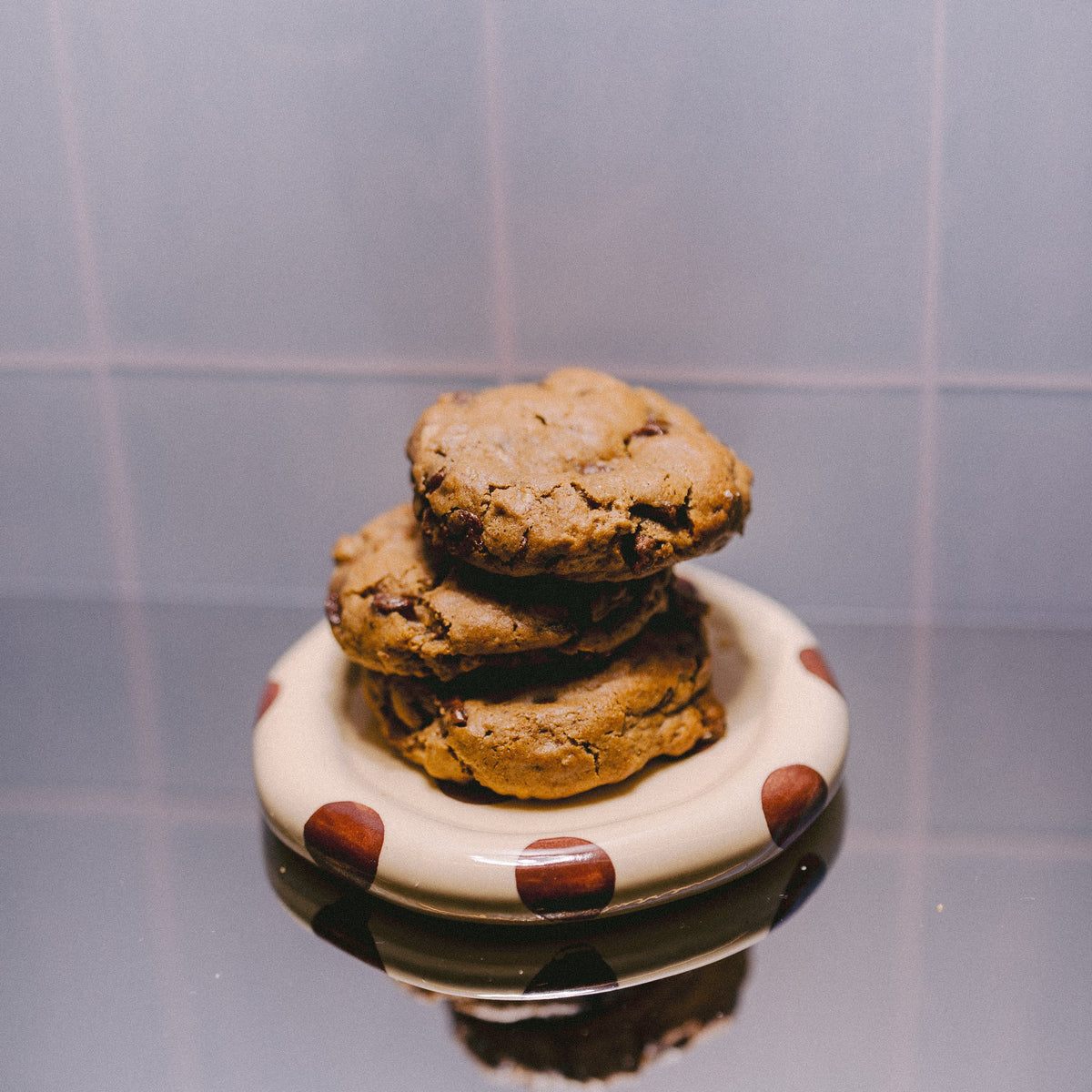 Three stacked freshly baked Milk it Classic chocolate chunk lactation cookies on a cream plate with brown polka dots, set against a blue tiled background with a reflective surface