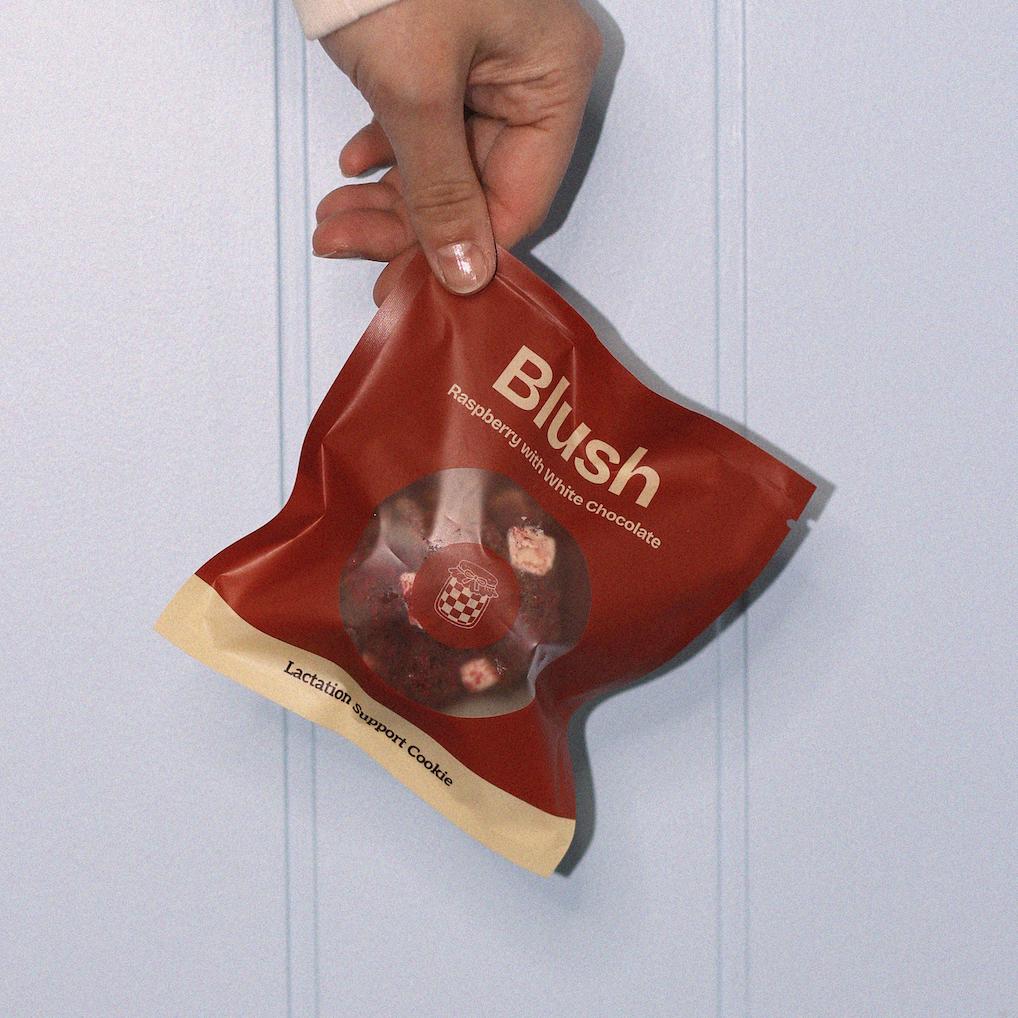 Hand holding a brown lactation cookie bag labeled 'Blush' against a light blue background