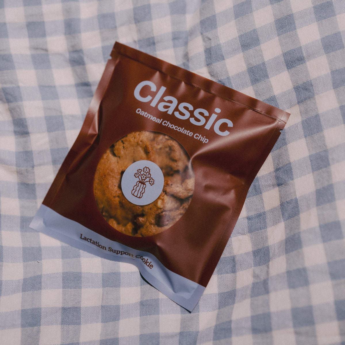 A bag of Milk it lacation support Classic Oatmeal Chocolate Chip cookies placed on a blue and white checkered blanket.