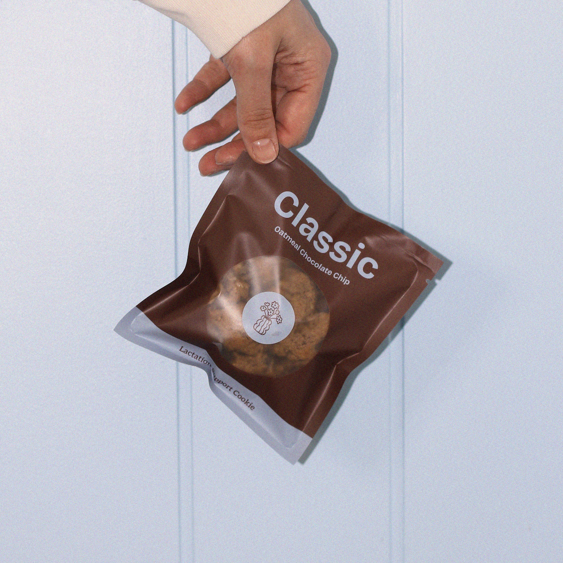 Hand holding a brown lactation cookie bag labeled 'Classic' against a light blue background