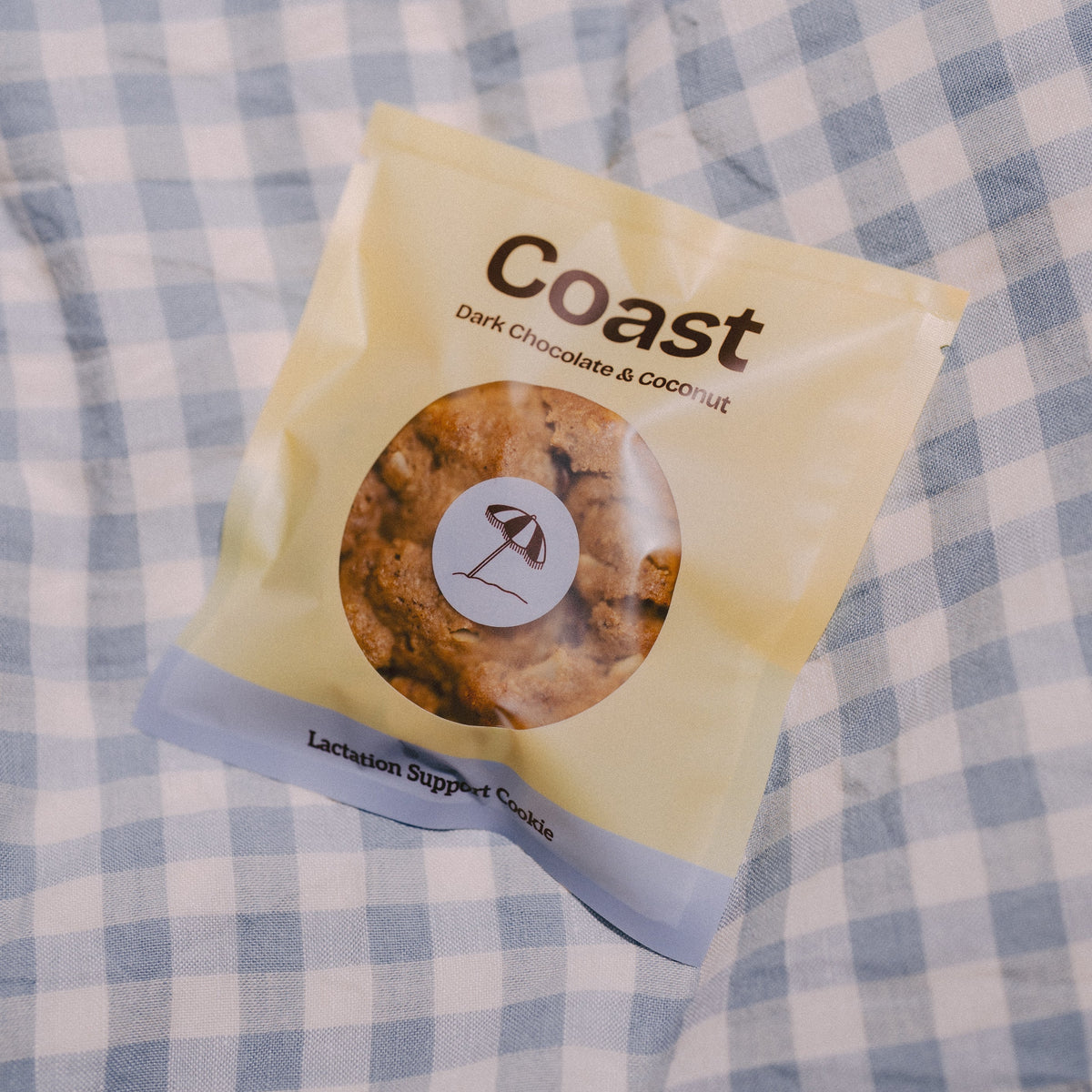 A single bag of Coast Dark Chocolate & Coconut Lactation Support Cookies lies on a blue and white checkered fabric surface.