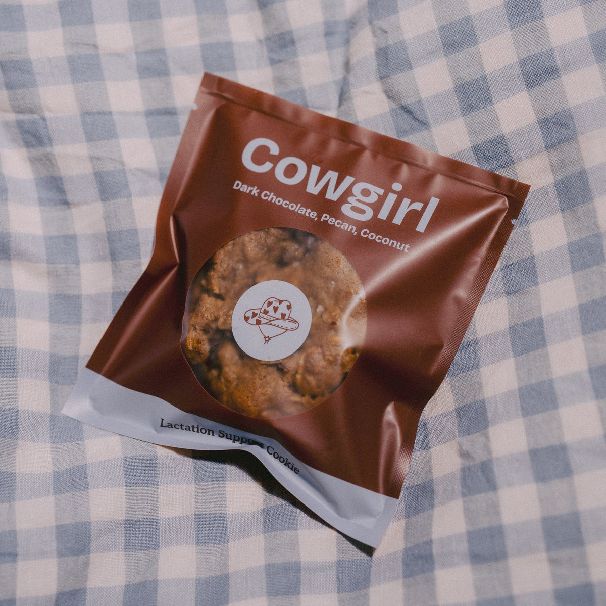 A bag of Milk it lactation support cookies in the flavour Cowgirl Dark Chocolate Pecon, Coconut cookies placed on a blue and white checkered blanket. The packaging is brown with white text and a photo of the cookie.