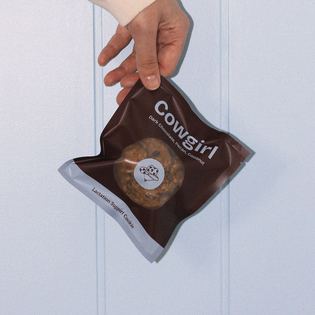 Hand holding a brown lactation cookie bag labeled 'Cowgirl' against a light blue background