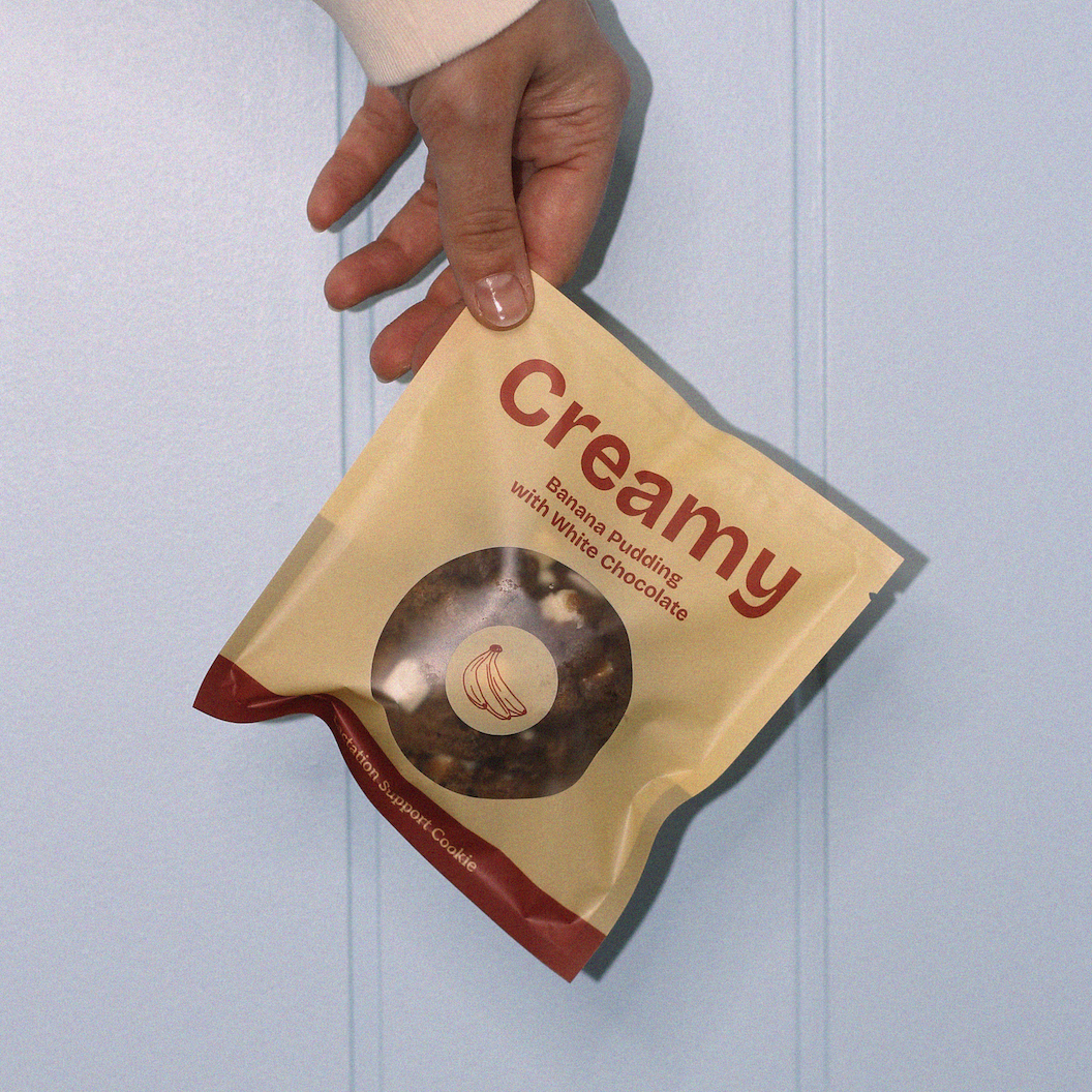 Hand holding a yellow lactation cookie bag labeled 'Creamy' against a light blue background