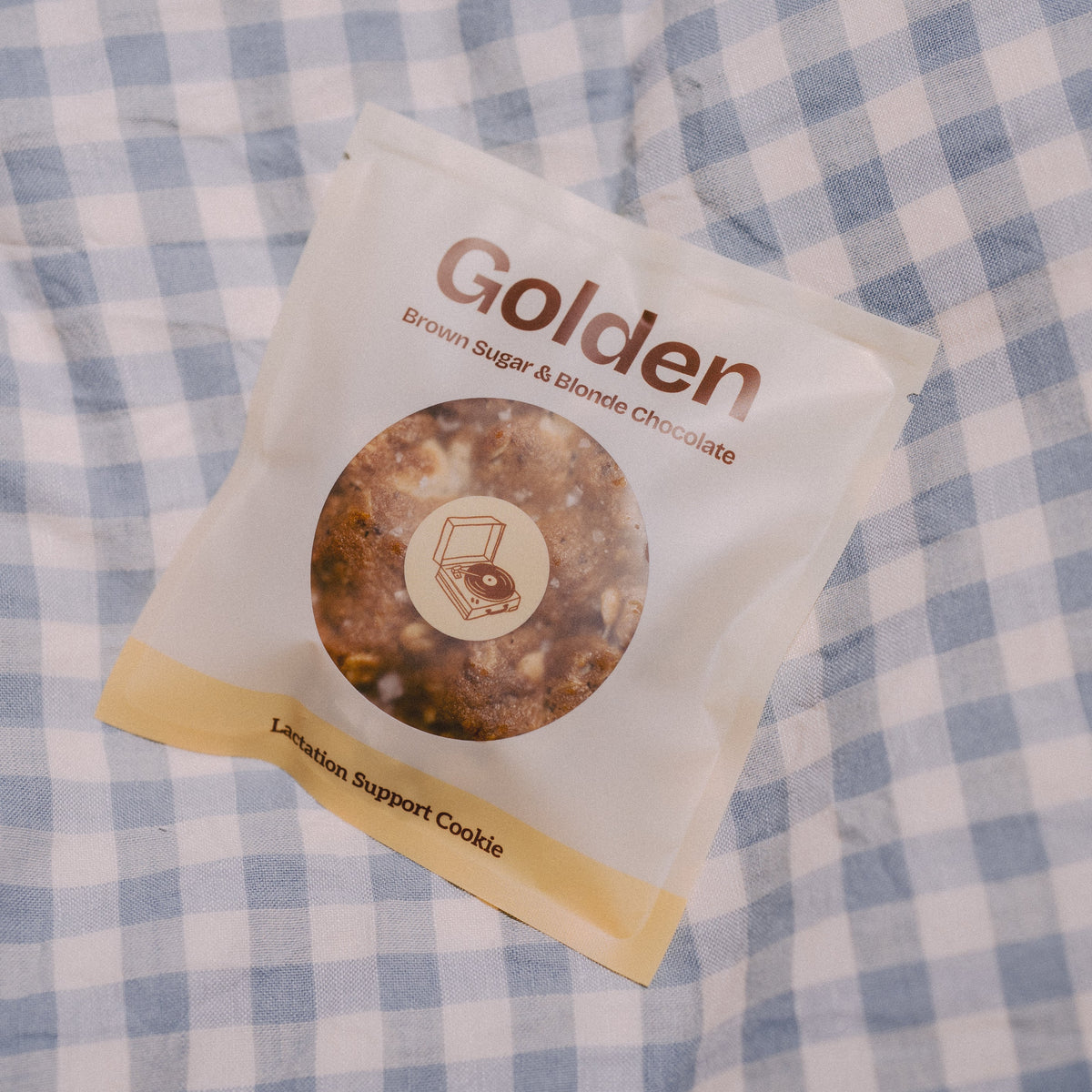 A single packaged Golden vegan lactation support cookie lies on a blue-and-white gingham fabric surface.