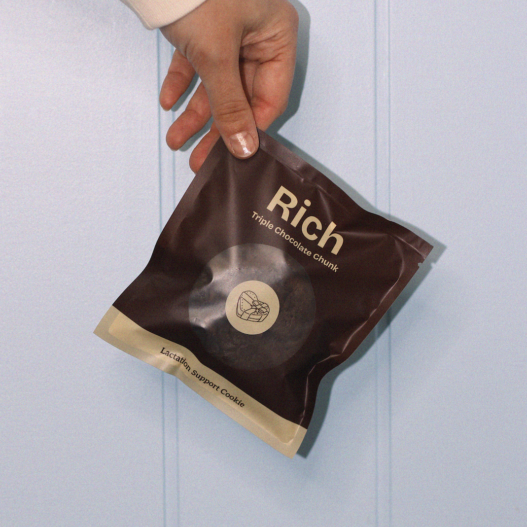 Hand holding a brown lactation cookie bag labeled 'Rich' against a light blue background