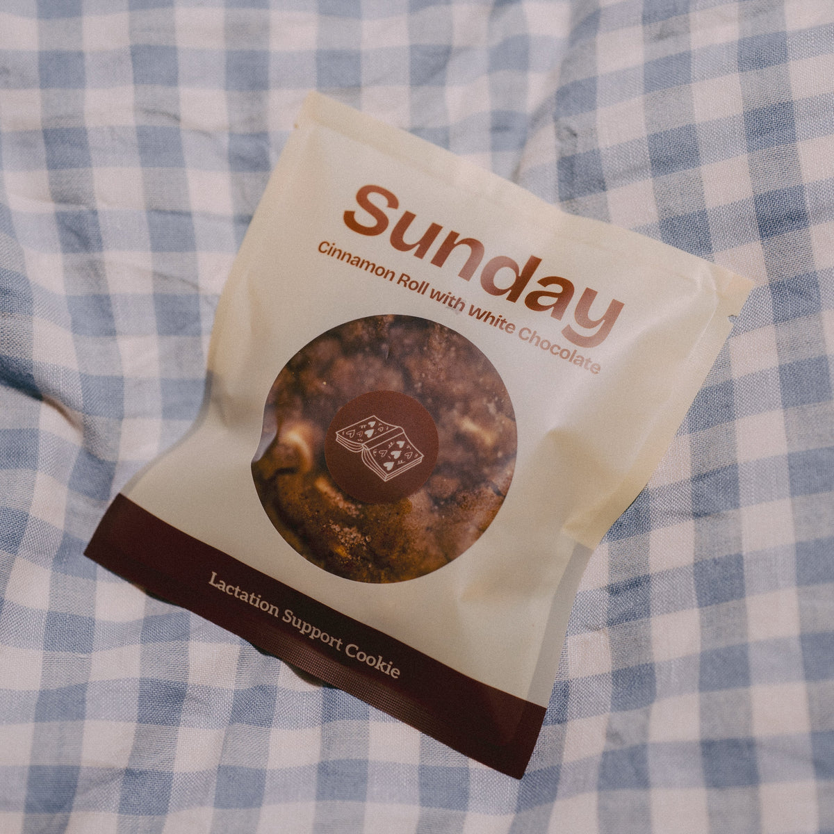 A bag of Sunday Cinnamon Roll with White Chocolate lactation support cookie resting on a blue and white checkered blanket.
