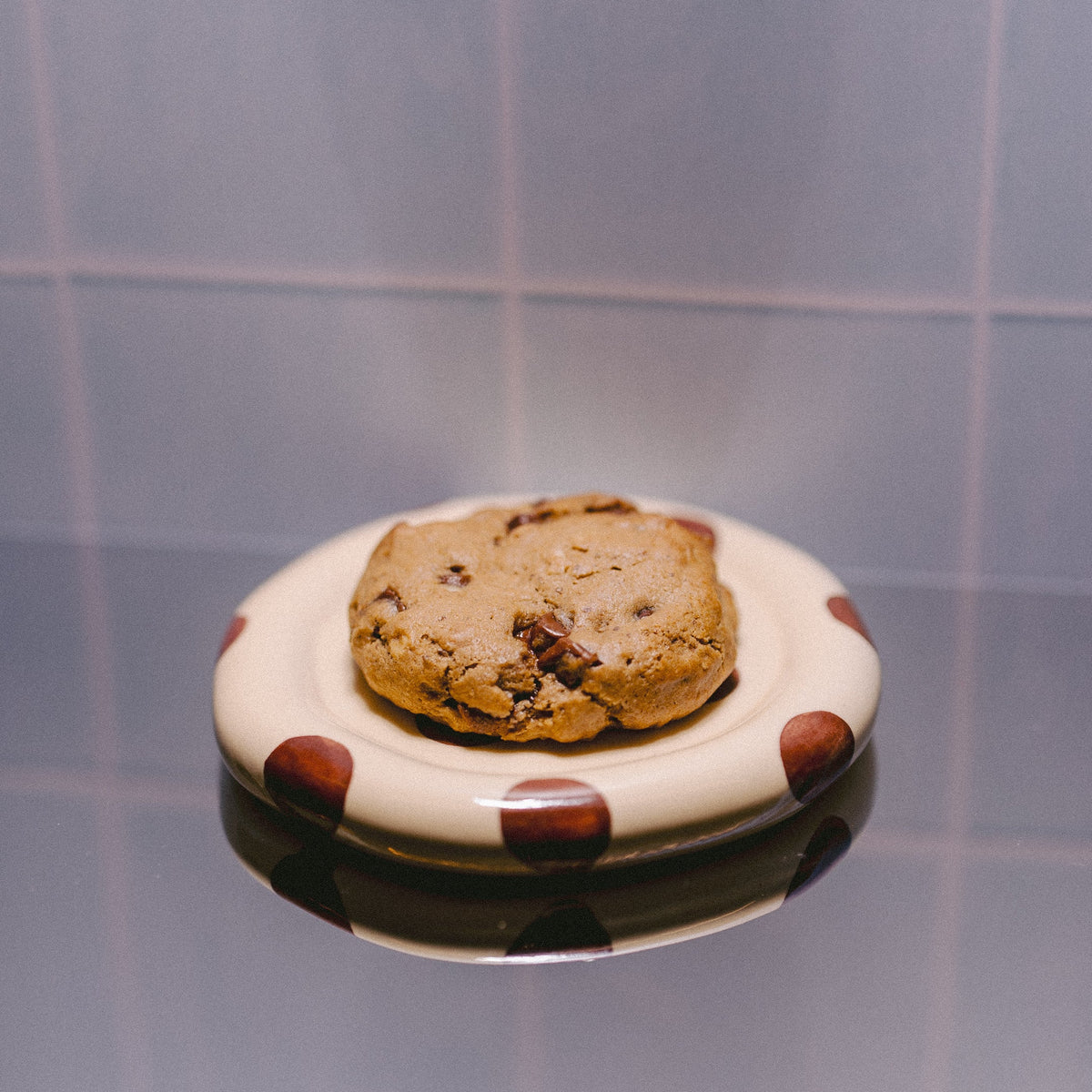 A single chocolate chip cookie sits on a small white plate with brown polka dots, set against a tiled background with a reflective surface below.