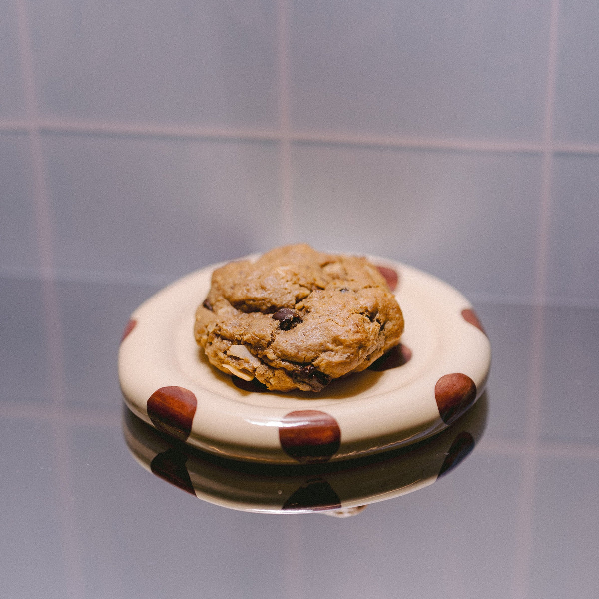 A single baked cookie sits centered on a small round white plate with brown polka dots, placed on a reflective surface against a blue tiled background.