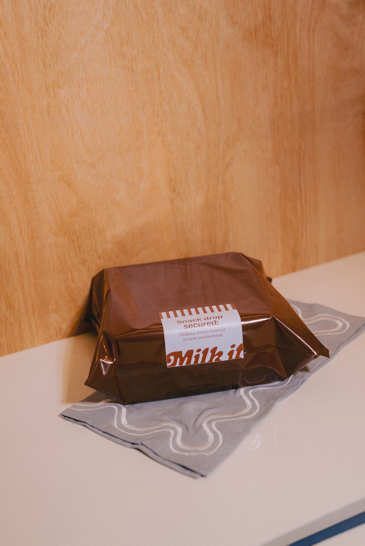 A brown wrapped packaged box of lactation support cookies with a blue label reading 'Milk it' sits on a blue patterned cloth against a light wood wall and white tabletop.