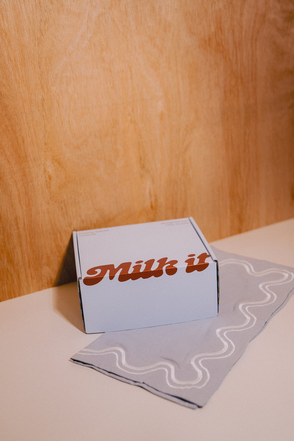 A blue box of lactation support cookies  with the words 'Milk it' in brown script sits on a light blue cloth with a wavy pattern, set against a warm wood-paneled background.