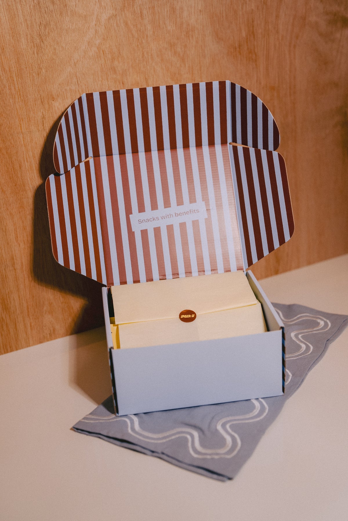 An open striped cardboard gift box of lactation support cookies with a cream-colored tissue, placed on a light blue patterned cloth against a wooden background.