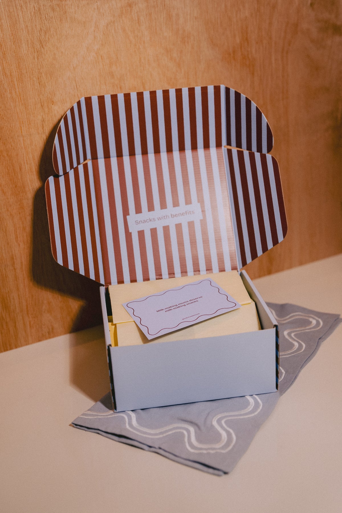 An open gift box with a brown and white striped lid, containing a small blue card, placed on a blue patterned cloth against a warm wood background.
