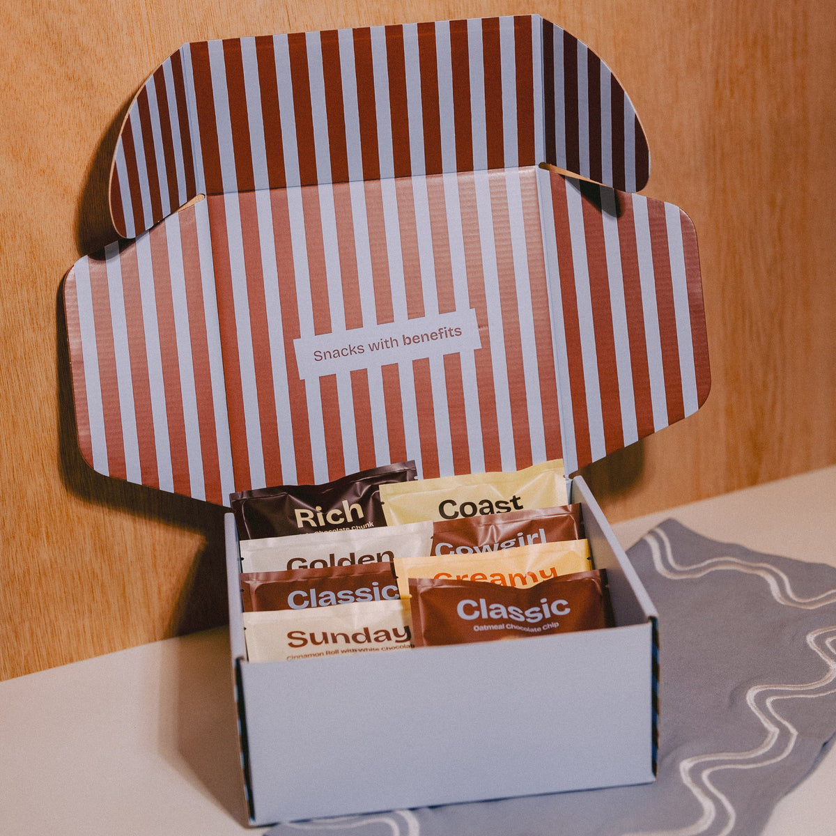 An open gift box of gourmet lactation support cookies displayed on a light surface against a wooden background. The box contains individually wrapped cookies labeled Rich, Coast, Golden, Sunday, and Classic, with a small card reading 'Snacks with benefits'
