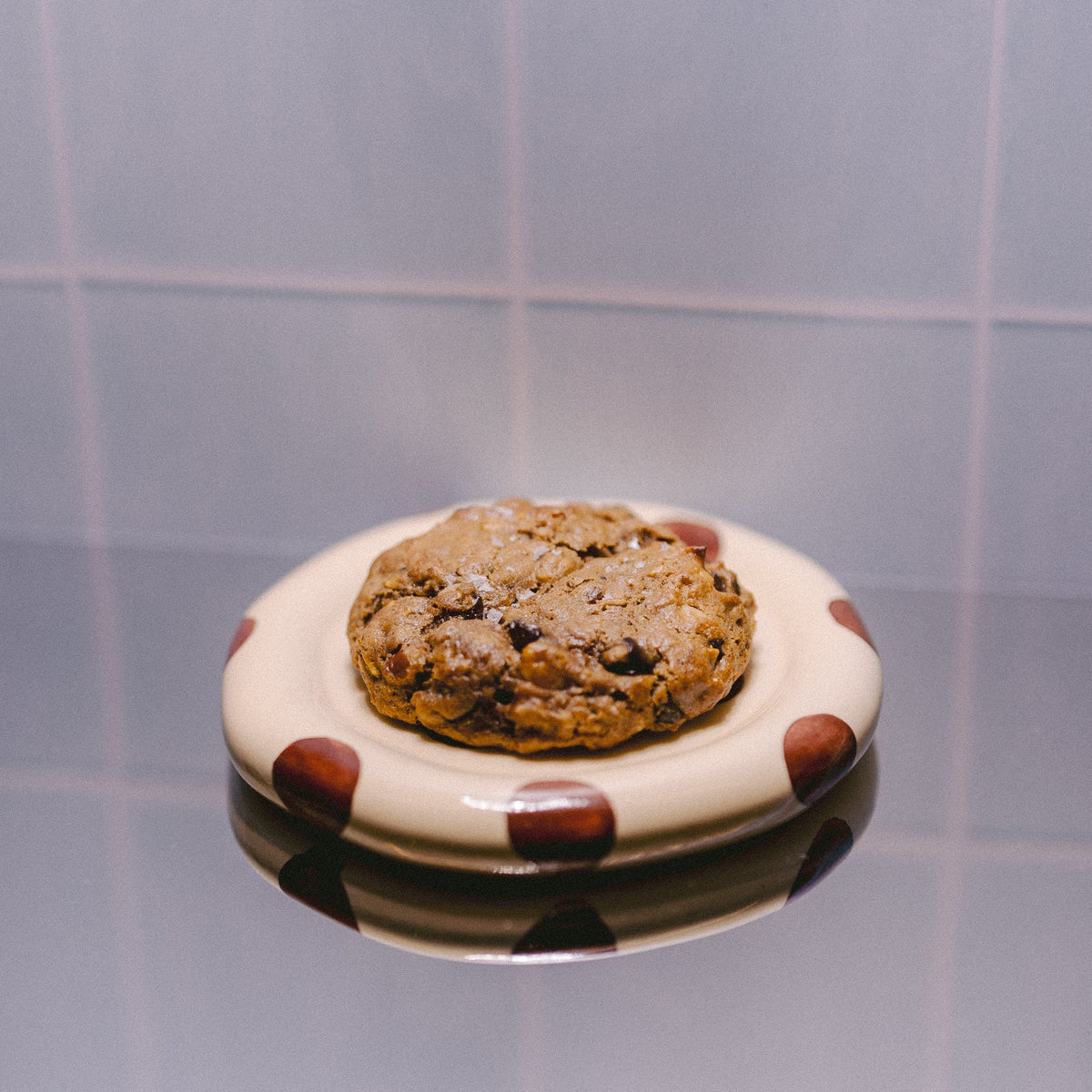 A dark chocolate, pecan, coconut and cornflakes cookie sits on a small white plate with brown polka dots, placed on a reflective surface against a light blue tiled background.