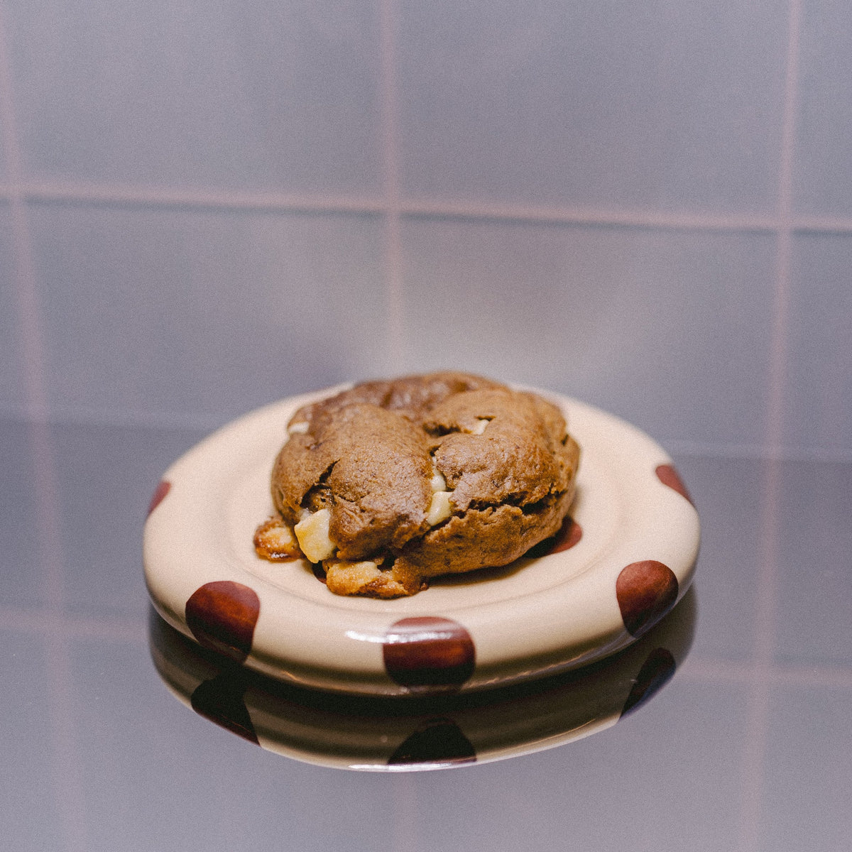 A single banana pudding with white chocolate cookie sits on a small white plate with brown polka dots, set against a tiled background with a visible reflection below.