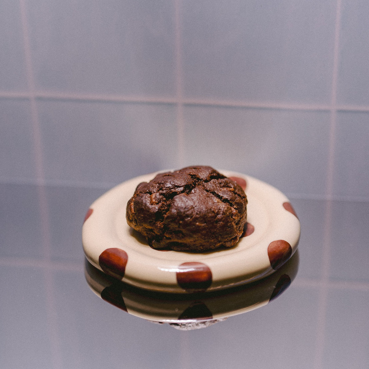 A small round white plate with brown polka dots holds a triple chocolate chunk baked lactation support cookie, set against a tiled background with a reflective surface below.
