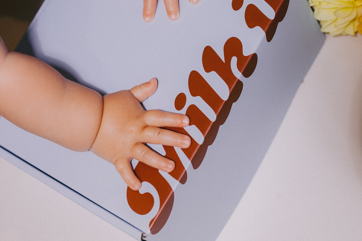A close-up lifestyle shot of a baby's hand resting on a blue Milk it box printed with the word 'Milk it' in large brown letters, with a baby arm also visible.