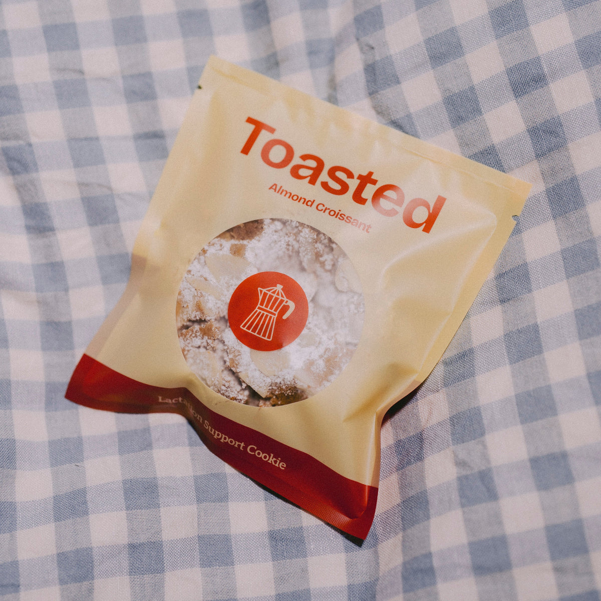 A bag of Toasted Almond Croissant lactation support cookies placed on a blue and white checkered blanket. The packaging is cream-colored with red text and a red bottom accent, featuring a circular window showing the cookies inside.