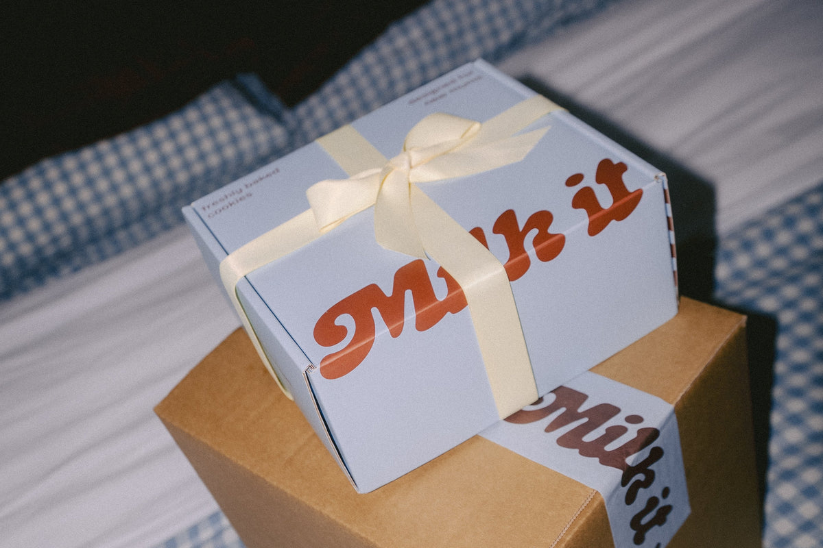 A blue gift box with brown 'Milk it' lettering and a cream ribbon sits on top of a larger brown cardboard shipping box on a blue-and-white checkered bed. The word 'Milk it' is also visible on a tape strip.