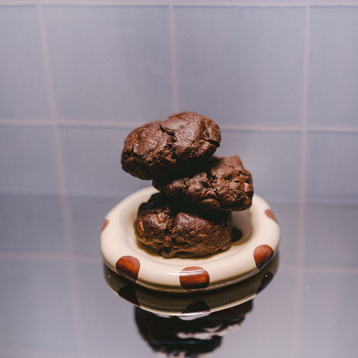 Three stacked freshly baked Milk it Rich triple chocolate chunk lactation cookies on a small cream ceramic dish with brown polka dots, set against a blue tiled background.