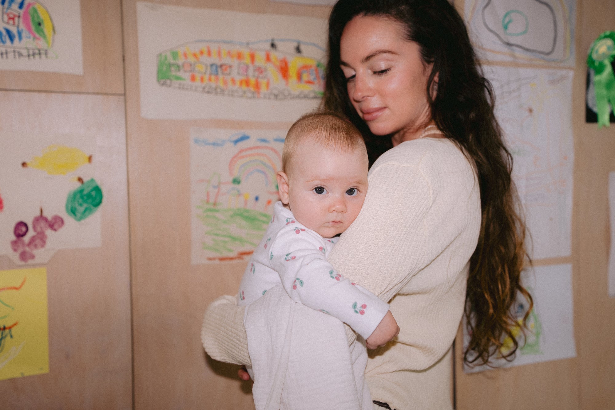 Mother holding her nine-month-old baby who rests her head on her shoulder, with kids’ artwork in the background