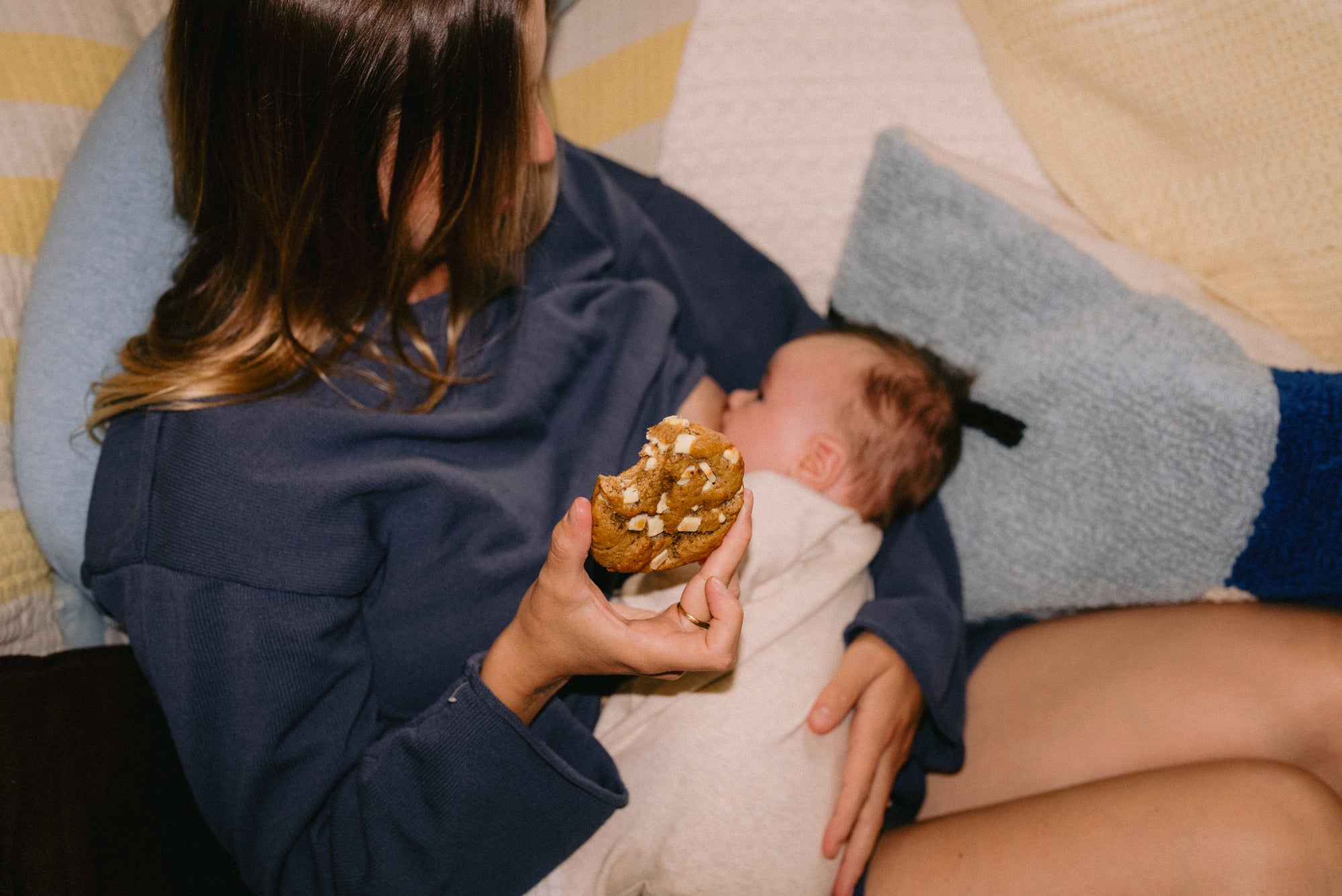 A new mum breastfeeding her six-week-old baby on the sofa, holding a Milk it cookie in one hand and gazing down lovingly at her daughter.