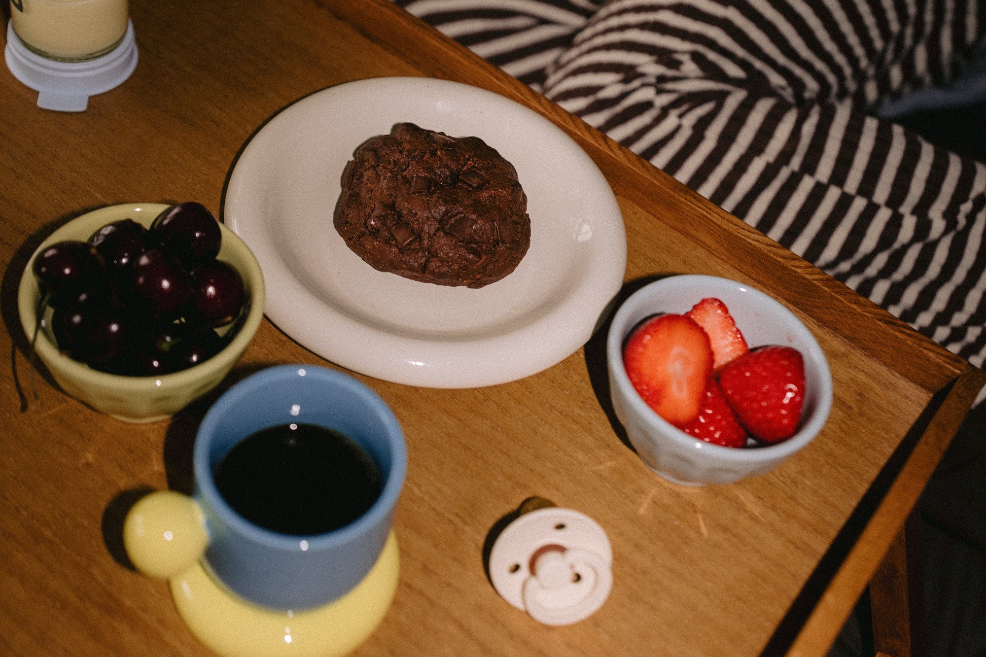 Rich triple chocolate lactation cookie on a white plate with black coffee, strawberries, cherries, and a dummy on a tray — the perfect breastfeeding snack moment.