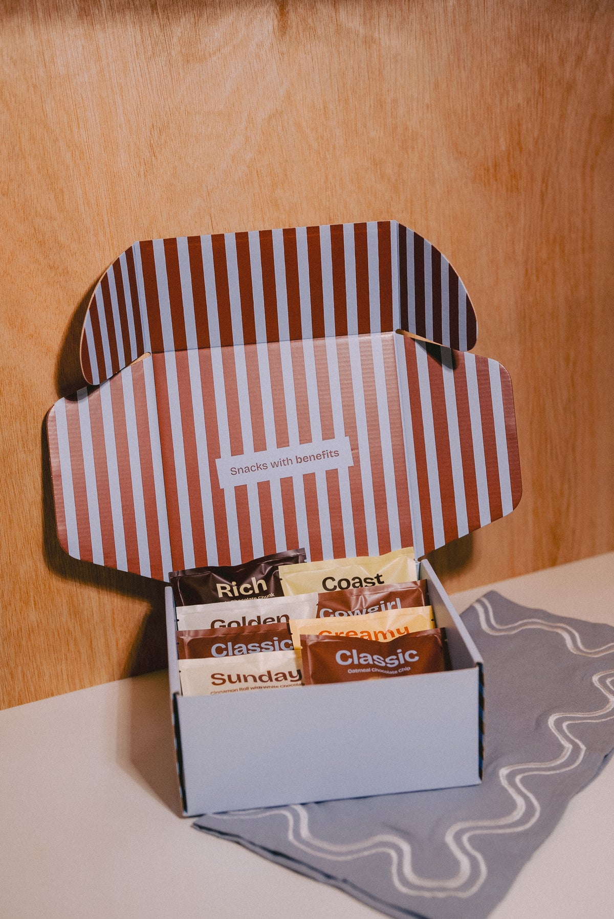 An open gift box of gourmet snacks with a brown and blue striped lid, displayed on a light surface against a wooden background. The box contains individually wrapped lactatioon support cookies labeled Rich, Coast, Golden, Sunday, and Classic, with a small card reading 'Snacks with benefits'.