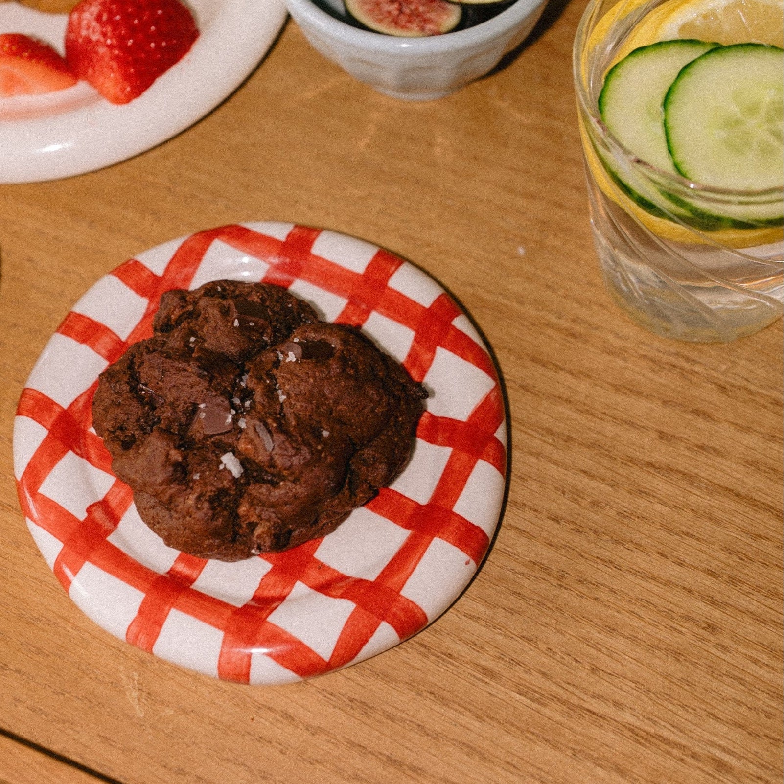 Triple chocolate chunk lactation cookie with flaky sea salt on a red and white gingham plate, styled with lemon cucumber water, figs, and strawberries