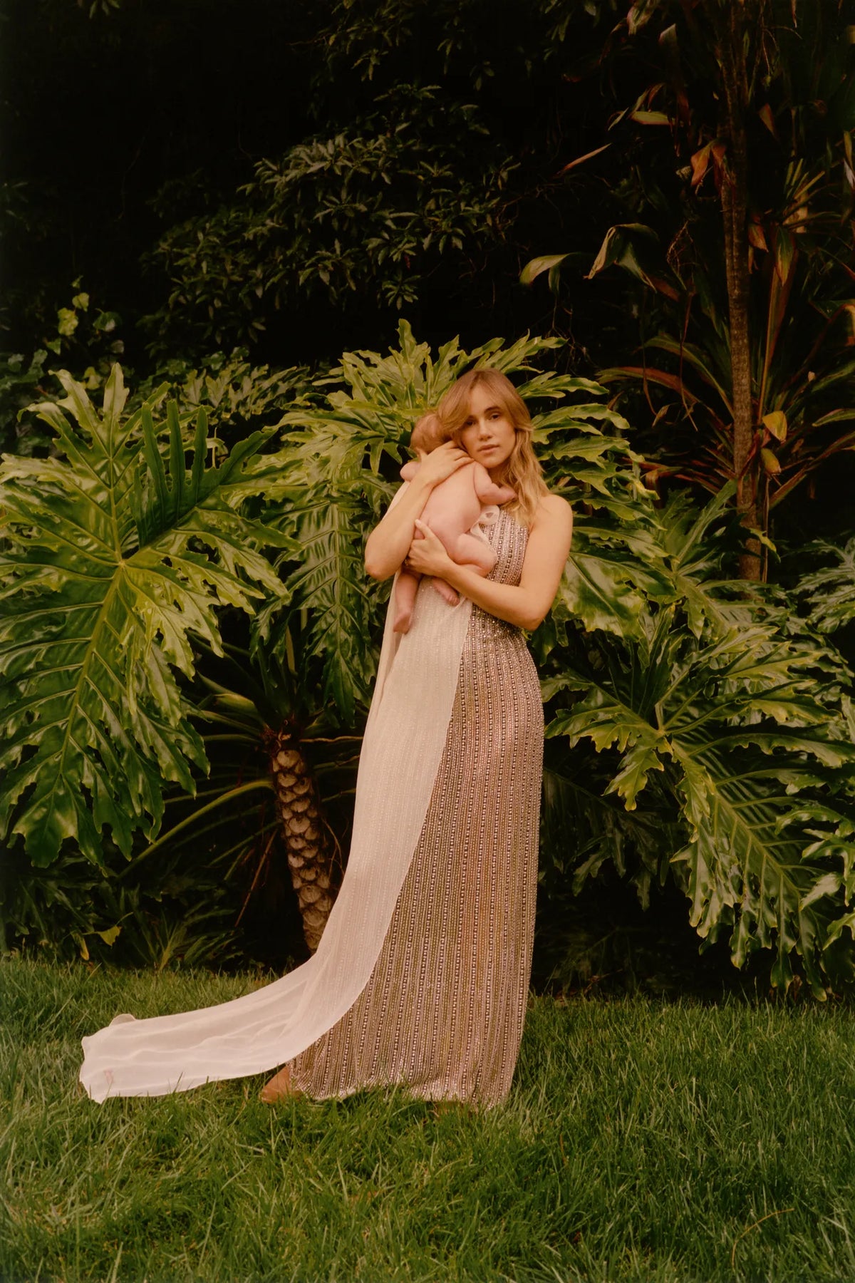 A woman in a shimmering pale pink sleeveless maxi dress stands on grass in a garden, holding a baby. Large green tropical plants fill the background.